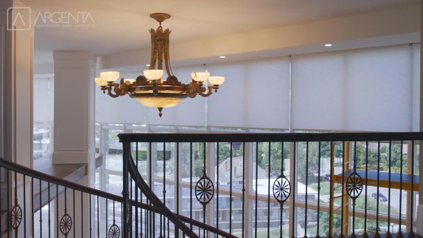 An elegant chandelier with glowing lights hanging over a modern staircase with large windows and semi-closed motorized shading.