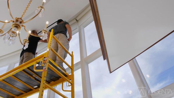 Two workers on scaffolding installing motorized shades near a chandelier.