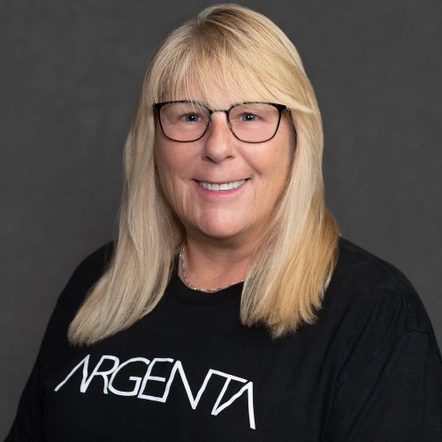 Portrait of a smiling woman with straight blond hair and glasses wearing a black Argenta T-shirt against a dark gray studio background.