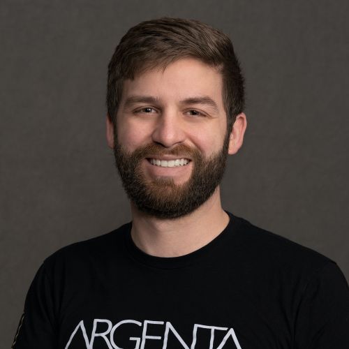 Portrait of a smiling man with short brown hair and a full beard wearing a black Argenta T-shirt against a dark gray studio background.