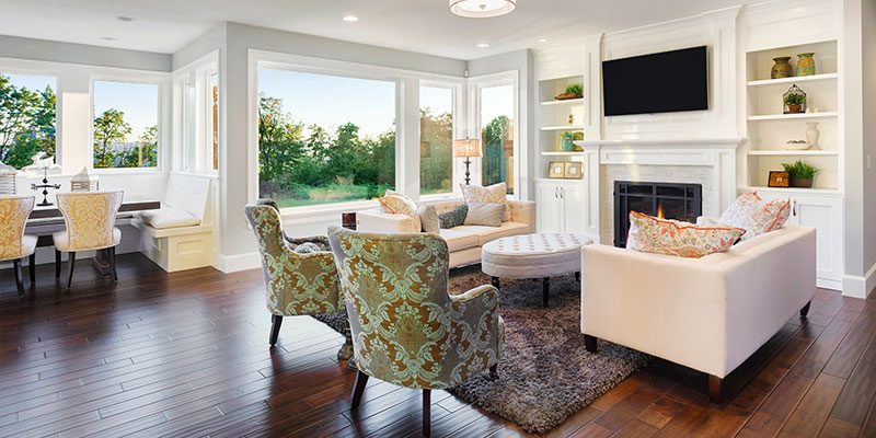Bright, open living room with large windows, white built-in shelves and fireplace, light-colored sofas, patterned armchairs around a round ottoman, and a dining nook with upholstered chairs on dark hardwood floors.