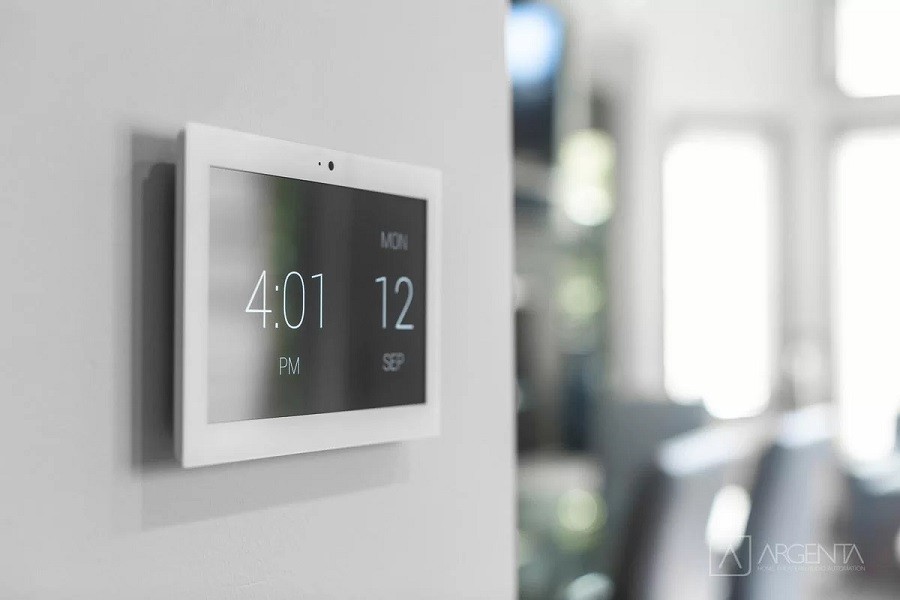 A sleek and sophisticated wall-mounted smart home touch screen frames the entrance to a well-lit modern living room. 