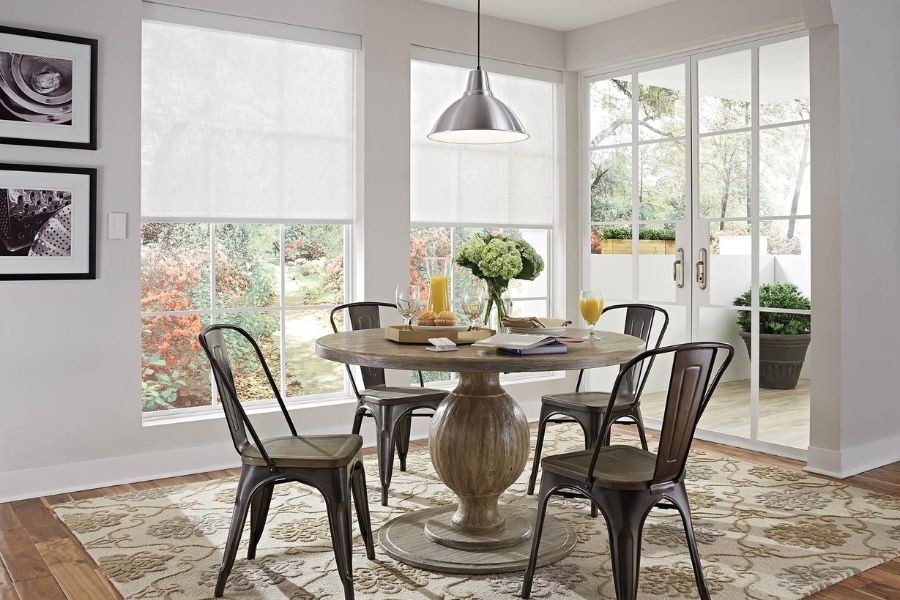 A round kitchen table adorned with plates and flowers, windows with motorized shades in the background.