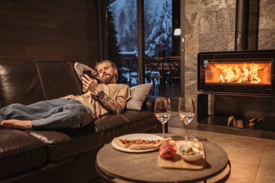  Man laying on the couch of his home next to a fireplace, looking at his phone, snow on the landscape beyond. 