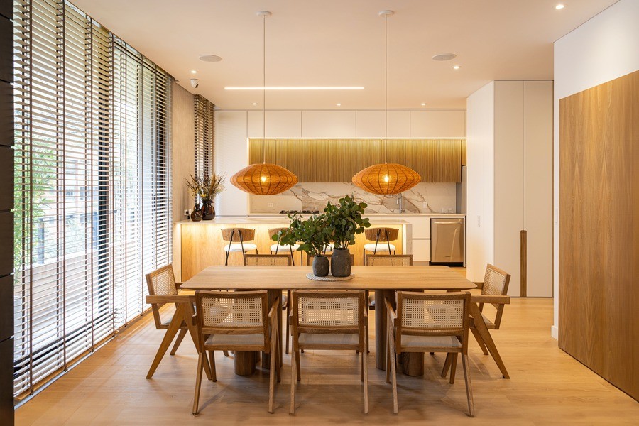 Modern interior design with rattan chairs sat at a kitchen table, with a well-lit kitchen in the background and rows of windows to the left. 