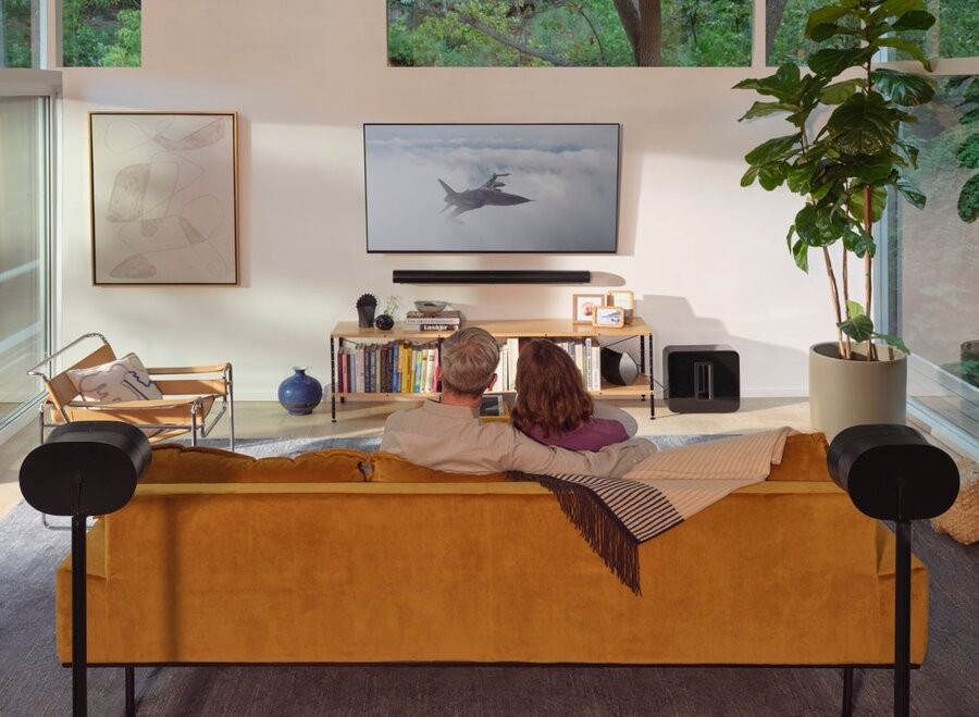 A media room with a couple sitting on the couch facing the TV with various Sonos multi-room music speakers surrounding them.