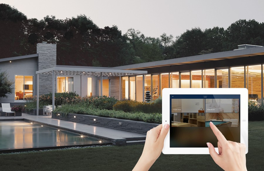 Exterior of a modern home at night, overlaid by hands holding a tablet with the Savant smart home interface.