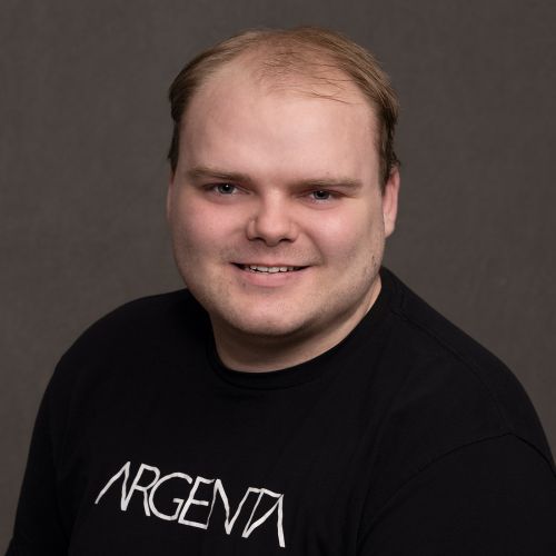 Headshot of a smiling man with short light brown hair wearing a black T-shirt against a gray background.