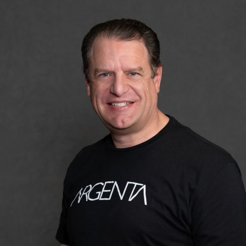 Headshot of a smiling man with slicked-back dark hair wearing a black T-shirt against a gray background.