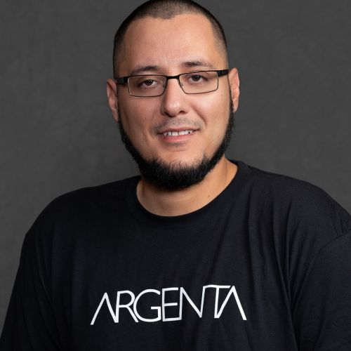 Portrait of a smiling man with short dark hair, glasses, and a goatee wearing a black Argenta T-shirt against a dark gray studio background.